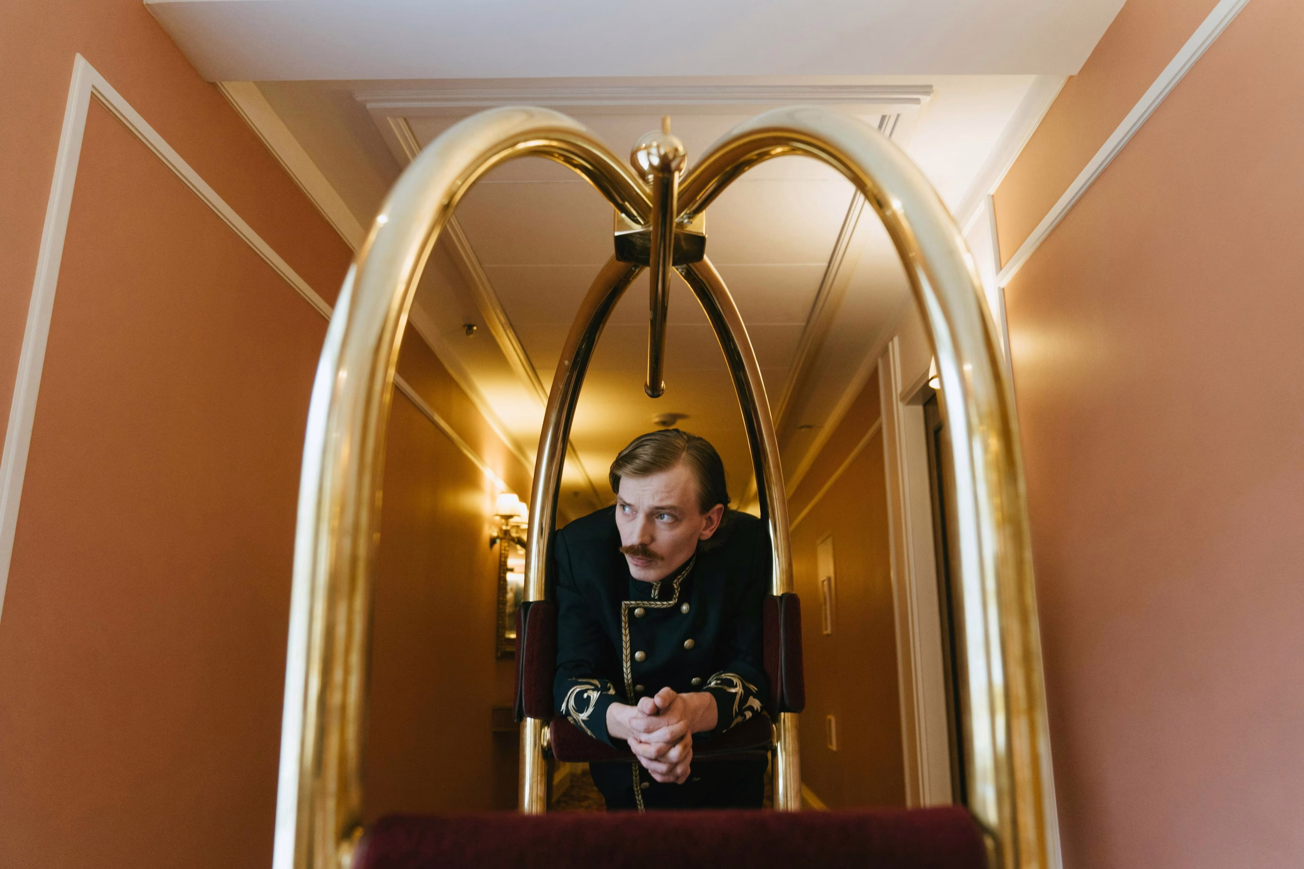 pexels-photo-5371575-5371575 Hotel bellhop dressed in a vintage uniform leaning on a luggage trolley in a brightly lit hallway.